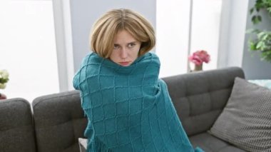 Sarı saçlı, mavi gözlü, düşünceli genç bir kadın. Gri bir kanepede turkuaz bir battaniyeye sarılı.