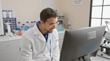 Laboratuvar ortamında laboratuvar önlüğü giymiş, sakallı, gülümseyen genç İspanyol bir adam bilgisayar ekranına bakıyor..