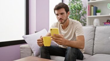 Sakallı genç bir İspanyol adam, elinde fincanla, rahat bir oturma odasındaki kanepede oturuyor.