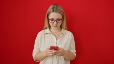 Neşeli genç sarışın bayan kırmızı duvarın üzerinden dostça bir mesajın keyfini çiviliyor. Dişlek bir gülümseme ve akıllı bir telefonla kendine güvenen bir şekilde duruyor.