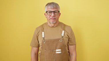 Endişeli orta yaşlı bir adam, gri saçlı ve gözlüklü, izole edilmiş sarı arka plana karşı önlük giymiş, gergin bir şekilde bir eli çenesinde, kemiren bir soruyu düşünüyor..