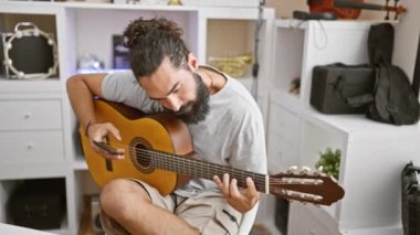 Sakallı İspanyol bir adam gitar çalıyor ve modern bir müzik stüdyosunda akıllı telefon kullanıyor.