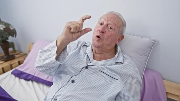 身穿睡衣的自信的老人用手指做着小动作 坐在床上的房间里 卧室尺寸概念展示小尺寸标志 — 图库视频影像