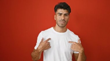 Beyaz tişörtlü, kendine güveni tam genç bir İspanyol, kırmızı arka planda poz veriyor.