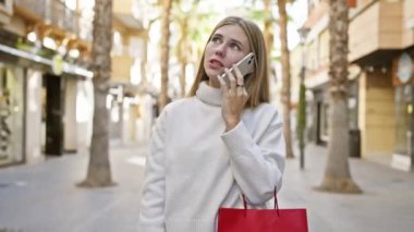 Sarışın, mavi gözlü bir kadın, güneşli bir caddede kırmızı bir alışveriş çantası taşırken akıllı bir telefonla konuşuyor..