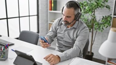 Modern bir ofis ortamında akıllı telefon ve tablet kullanan kulaklık takan olgun sakallı bir adam..