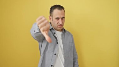 Kızgın görünüşlü, tişörtlü genç İspanyol bir adam, olumsuz bir jestle baş parmağını kaldırıyor, izole edilmiş sarı arka planda mutsuz bir ifade sergiliyor.