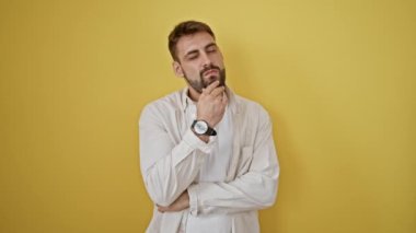 Düşünceli genç İspanyol bir adam, sakin bir görünüm sergiliyor, izole edilmiş sarı bir arka plana karşı duruyor, şüpheli bir ifade yayıyor, ama kurnaz bir fikri ateşliyor gibi görünüyor..