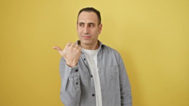 İzole edilmiş, güneşli, sarı bir duvarın önünde dikilen, tişörtlü, neşeli, genç İspanyol bir adam. Neşeyle işaret ediyor ve yana bakıyor, başparmağını kaldırıyor, bulaşıcı bir gülümseme sergiliyor..