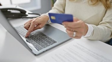 Yetişkin bir kadının elleri dizüstü bilgisayar kullanırken ve bir ofis ortamında kredi kartı tutarken..