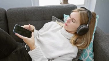 Kulaklıklı genç bir kadın akıllı telefon kullanarak kanepede dinleniyor, rahat bir oturma odasında boş zamanlarını özetliyor..