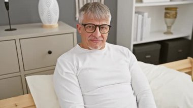 Pijamalarıyla gülen neşeli orta yaşlı bir adam, neşeli bir dille konuşuyor, zafer işaretlerini güneşli yatak odasında sergiliyor..