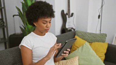 Gülümseyen Afrikalı Amerikalı kadın evdeki kanepede oturuyor, rastgele tablet kullanıyor..