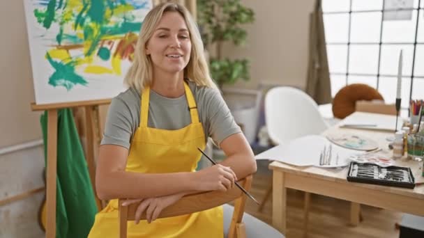 一个穿着黄色围裙 拿着画笔在艺术工作室里微笑的女艺术家 — 图库视频影像
