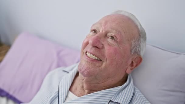 一个快乐的老人 坐在床上 充满自信地陷入沉思 面带微笑地离去 睡衣中一幅经典的室内肖像画 放在舒适的卧室里看一边 — 图库视频影像