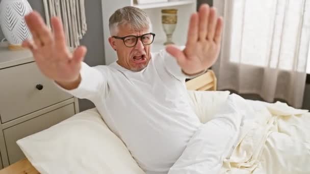 パジャマの中年の灰色の髪の男性は 彼の居心地の良い寝室で怒りの明確な表現を披露し 彼の手の決定的な停止ジェスチャーにイライラを表しています — ストック動画