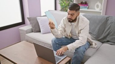 Sakallı bir adam, modern bir apartmanın oturma odasında dizüstü bilgisayarıyla çalışırken kendini bir vantilatörle serinletir..