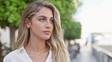 Havalı, rahat genç sarışın kadın ciddi ciddi kameraya bakıyor, güneşli şehir sokağında gelişigüzel duruyor, çekici bir yaşam tarzı portresi var, şehir arka planında.