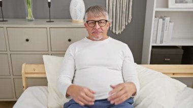 Pijama giymiş neşeli, orta yaşlı bir adam, rahat yatak odasında bir karşılaştırma konsepti sunuyor, avuçlarını akıllıca dengeliyor, dokunaklı el hareketleriyle reklamını yaparken gülümsüyor..