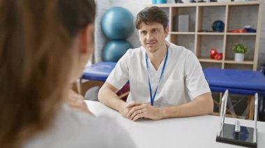 Beyaz önlüklü bir adam, iyi donanımlı bir fizyoterapi kliniğinde bir hastaya danışmanlık yapar. Rehabilitasyon topları ve tedavi yatağı sunar..