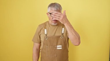 Şok olmuş gri saçlı, gözlüklü, orta yaşlı bir adam, bir önlük takıyor; dikizlerken yakalandı, utangaç yüzünü bir elle örtüyor, çekingen gözler açık sarı bir arka fona uzanmış parmaklarından dışarı bakıyor.