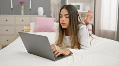 Beyaz tişörtlü genç bir İspanyol kadın modern bir yatak odasında rahat bir yatak ve doğal bir dekorla dizüstü bilgisayarı üzerinde çalışırken gülümsüyor..