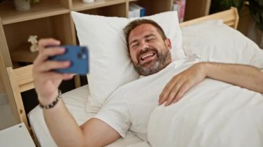 Yatakta uzanmış gülümseyen bir adam selfie çekerken sıcak bir odada bir anlık mutluluğu yakalar..