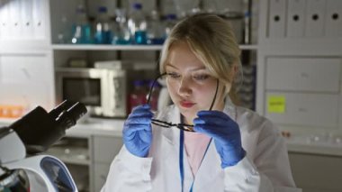 Sarışın kadın bilim adamı gözlüklerini laboratuvar ortamında ayarlıyor, ekipmanlarla çevrili..