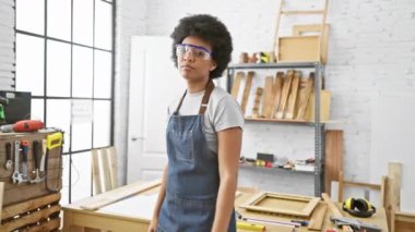 Kıvırcık saçlı, güvenlik gözlüğü takan ve kot önlüklü bir kadın iyi aydınlatılmış bir marangoz atölyesinde duruyor..