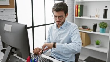 Gözlüklü ve sakallı, açık mavi gömlekli genç bir İspanyol, parlak bir ofiste çalışırken zamanı kontrol ediyor..