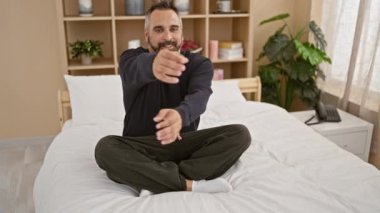 A bearded man with grey hair stretches while sitting cross-legged on a white bed in a well-lit bedroom, emanating a sense of wellness and comfort at home.