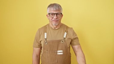 Neşeli, gri saçlı, orta yaşlı, gözlük ve önlük takan, şakacı, gözlerini şaşkına çeviren bir adam. Güneşli sarı bir arka planda izole edilmiş.