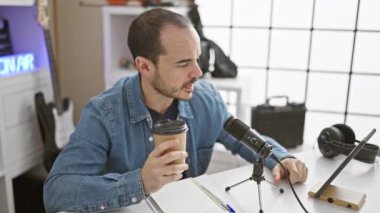 Kel ve sakallı bir adam içeride mikrofon ve tablet kullanarak podcast yaparken kahvesini yudumluyor..