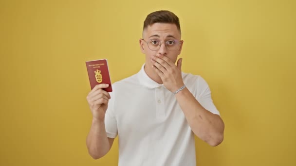 ショックを受けた若いヒスパニック系男性は手で口を覆い デンマークのパスポートを保持しています 沈黙のサプライズ 黄色の背景に孤立した重大な間違いの恐れ — ストック動画