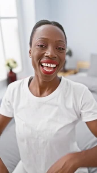 陽気なアフリカ系アメリカ人女性が屋内で遊び心のある瞬間を過ごし アパートのベッドに横たわり 喜びの表情を求め ピーク ガーを演奏し ベッドルームで笑顔を見せました — ストック動画