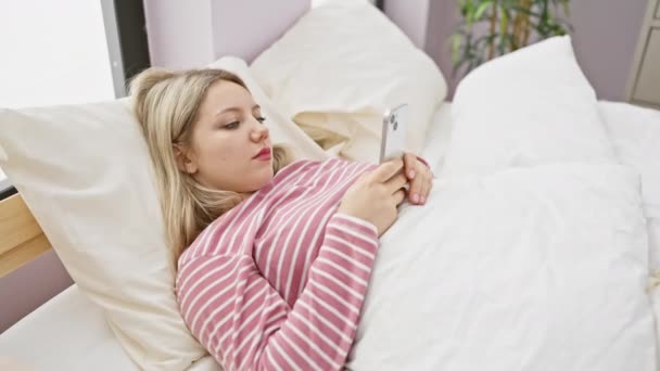 ストライプのシャツの金髪の女性は明るい寝室の設定で彼女のスマートフォンに従事ベッドにあります — ストック動画