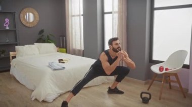 Sakallı bir adam ağırlıklar ve tarafsız bir dekorla yatak odasında egzersiz yapıyor, evde bir spor salonunun rutinini gösteriyor.