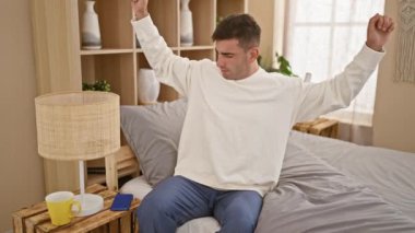 Yakışıklı, Latin asıllı, yatakta dinlenen, akıllı telefonunu rahat bir yatak odasında tutarken kollarını uzatan bir adam.