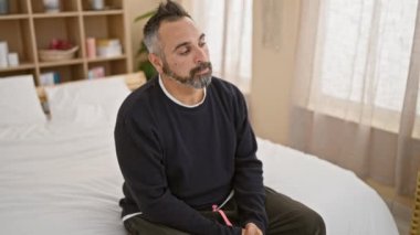 Düşünceli, olgun, gri sakallı bir İspanyol, yatak odasının içinde, yatağın üzerinde oturuyor.
