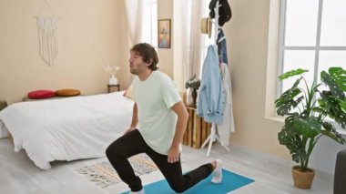 Genç bir adam yatak odasında yoga minderi, bitki ve yatak ile egzersiz yapıyor..