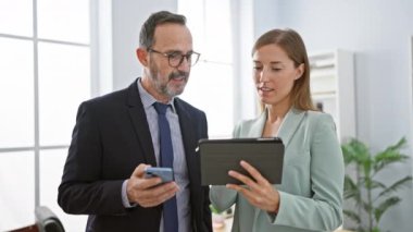 İki iş adamı gülümsüyor, erkek ve kadın, şık ofis içinde birlikte duruyorlar, dokunmatik dokunmatik ve akıllı telefonu ustalıkla kullanıyorlar çevrimiçi çalışırken