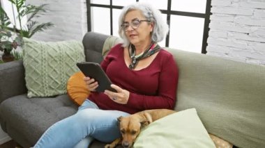Gri saçlı, gözlüklü ve kırmızı süveterli olgun bir kadın koltukta tablet kullanırken gülümsüyor ve evinde rahat bir köpeği var..
