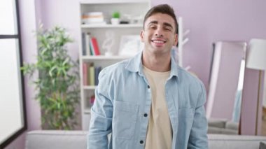 Evde kendine güvenen genç İspanyol bir adam, pozitif bir ifadeyle kanepede dinleniyor ve gülümsüyor, evin içinde rahat olmanın keyfini çıkarıyor.