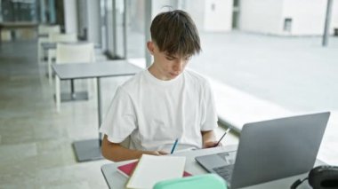 Modern üniversite kütüphanesinde dizüstü bilgisayar ve defterlerle çalışan beyaz bir genç.