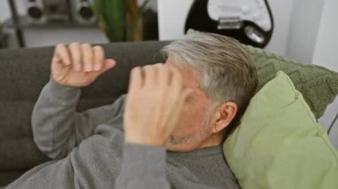 Evde yeşil yastıklı ve modern iç mimarisi olan bir koltukta dinlenen gri saçlı rahat bir son sınıf öğrencisi..