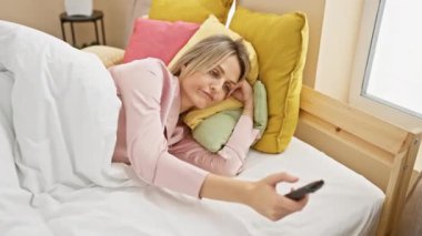 Pijamalı yorgun, beyaz bir kadın yatak odasında uzaktan kumandayla yatıyor..