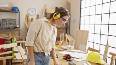 Genç bir esmer kadın, iyi ışıklandırılmış bir marangozluk stüdyosunda gülümserken güvenlik giysileri giyer, profesyonel bir marangozluk ortamı yayar..