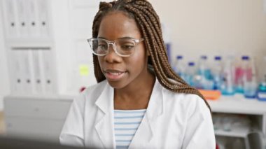 Gözlük takan odaklanmış Afrikalı bir kadın ve laboratuvar ortamında bilgisayar ekranına bakan laboratuvar önlüğü..