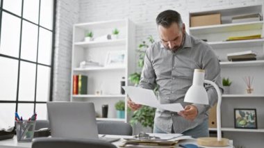 Sakallı, gri saçlı, modern ofis içinde gazete okuyan Latin kökenli olgun bir adam..