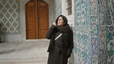 İstanbul 'un tarihi yerinde gözlüklü ve başörtülü gülümseyen kadın süslü fayansların yanında duruyor.